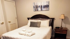 Un lit bien fait, avec une couette blanche et des oreillers, une table de chevet avec une lampe, et une porte sur la gauche.