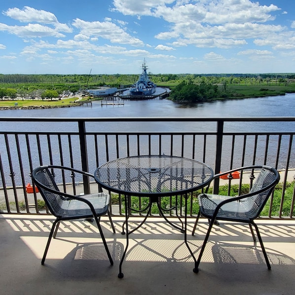 Ein Balkon mit einem runden Metalltisch und zwei Stühlen, mit Blick auf einen Fluss, auf der gegenüberliegenden Seite liegt ein Schiff am Anleger.
