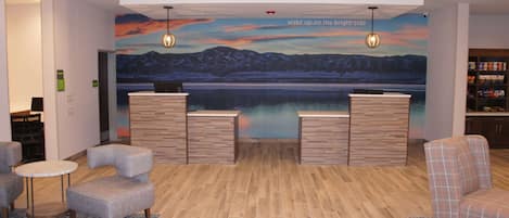 A reception area with a wooden reception desk, two armchairs, a small round table, and a mural of a lake with mountains.
