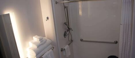 A modern bathroom with a glass-enclosed shower, a handrail, a towel rack with folded towels, and a small black chair.