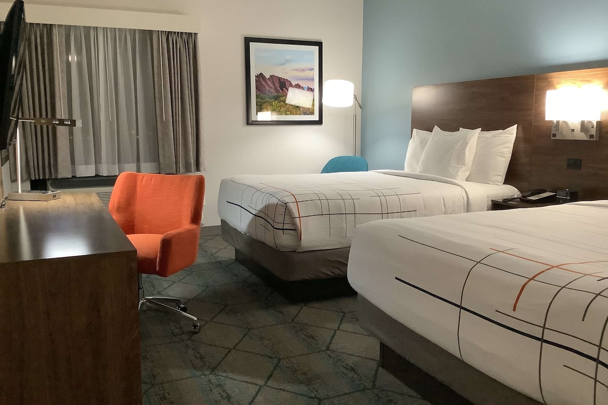 Habitación de hotel con dos camas, un escritorio, una silla, una lámpara y un cuadro en la pared.