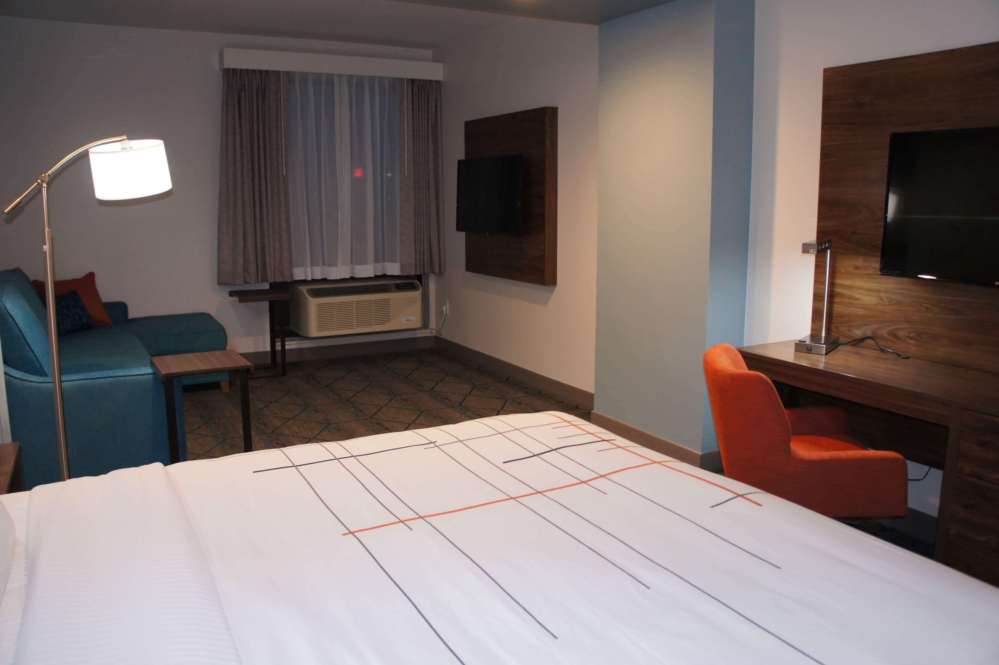 Una camera d'albergo con un letto, una scrivania, una sedia, una televisione, una lampada e una finestra con tende.