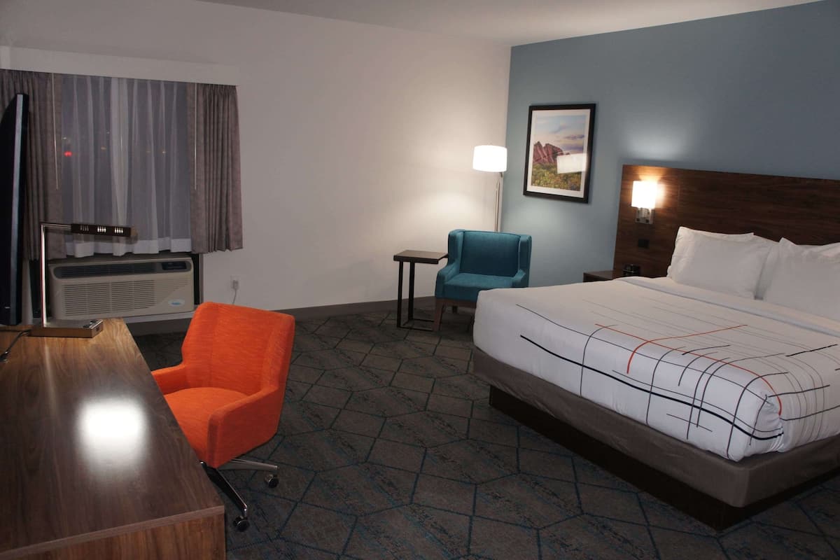 Habitación de hotel con una cama grande, un escritorio, una silla naranja, una mesita pequeña, una lámpara y un cuadro en la pared.