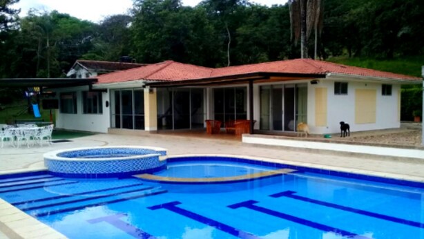 A swimming pool with a tiled deck, a covered patio area, and a house with a red roof in the background. - Casa con Piscina Privada Anapoima (Anapoima)