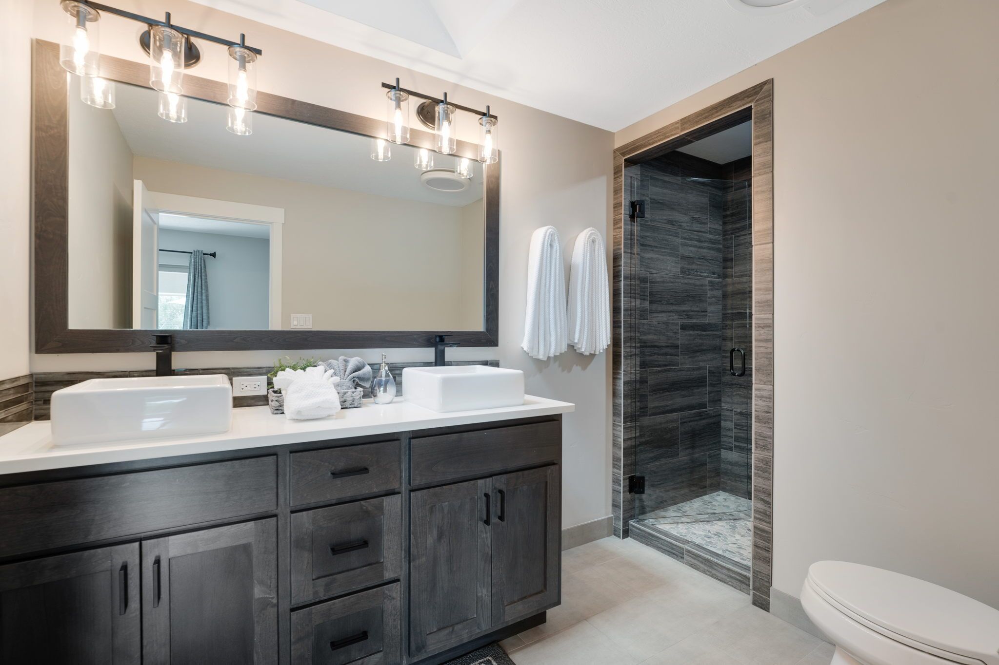 A modern bathroom with a large mirror, dual sinks, dark wood cabinets, and a glass-enclosed shower.