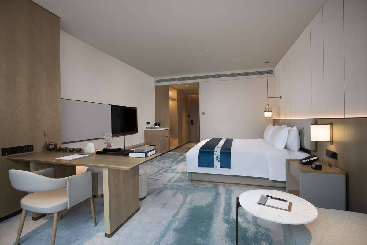 A hotel room with a bed, desk, chair, bedside table, and a TV.