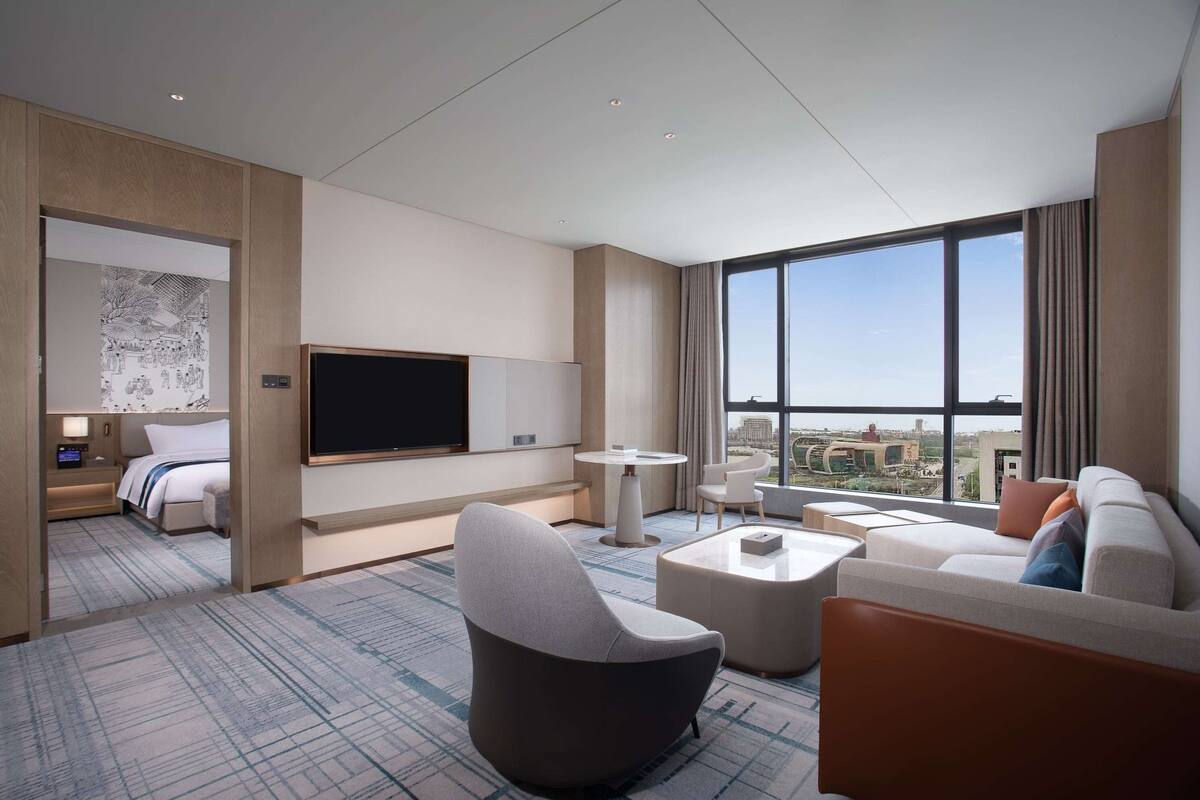 A modern hotel room with a large flat-screen TV, a comfortable sofa, a dining area, and a view of the city.