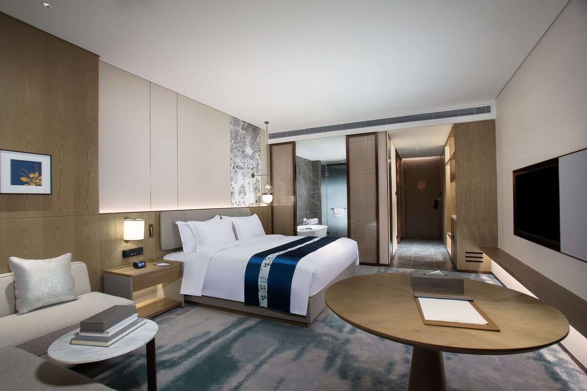 A modern hotel room with a neatly made bed, a bedside table with a lamp, a small round table with a notepad, and a TV mounted on the wall.
