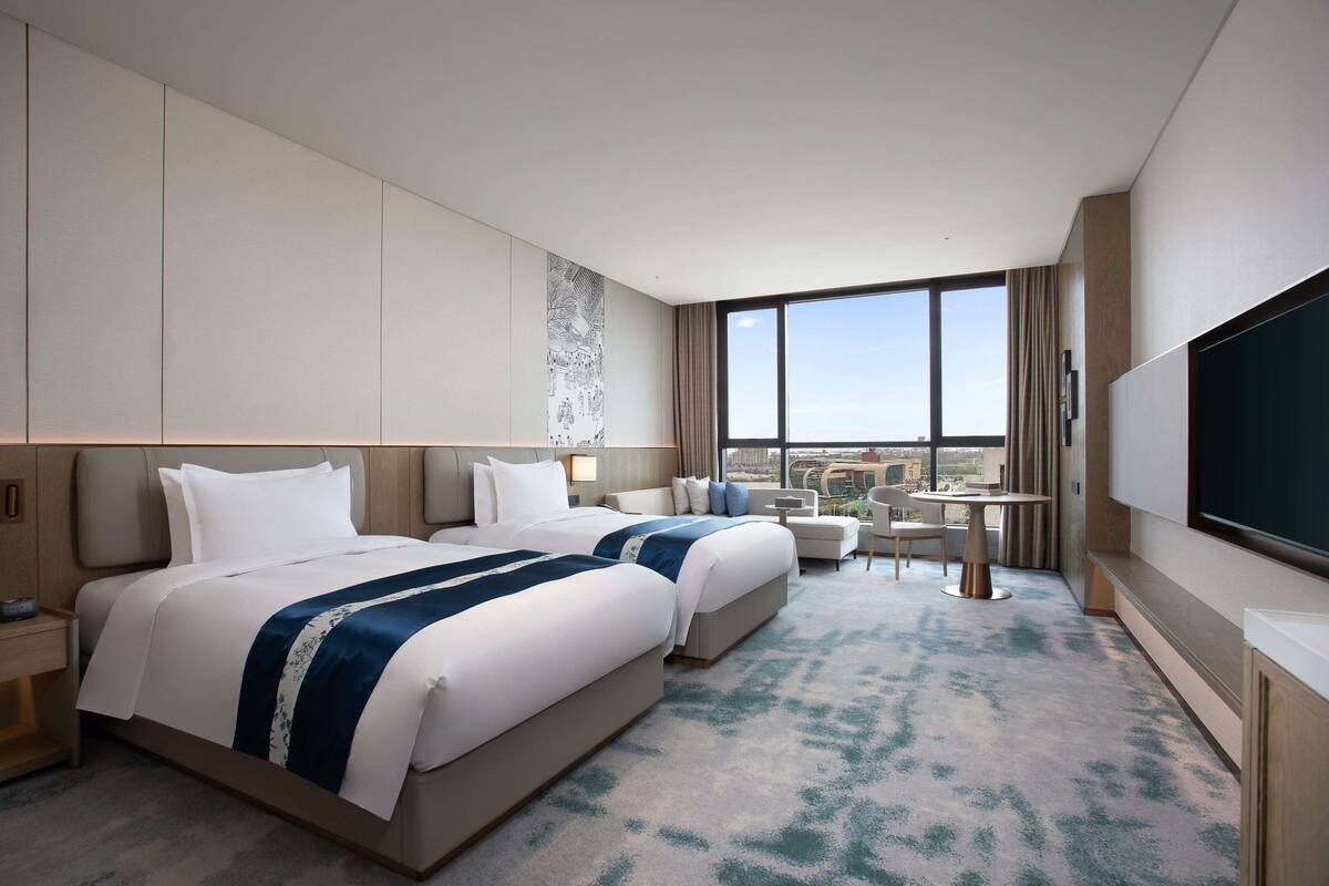 A hotel room with two beds, a desk, a chair, a TV, and a large window with a city view.