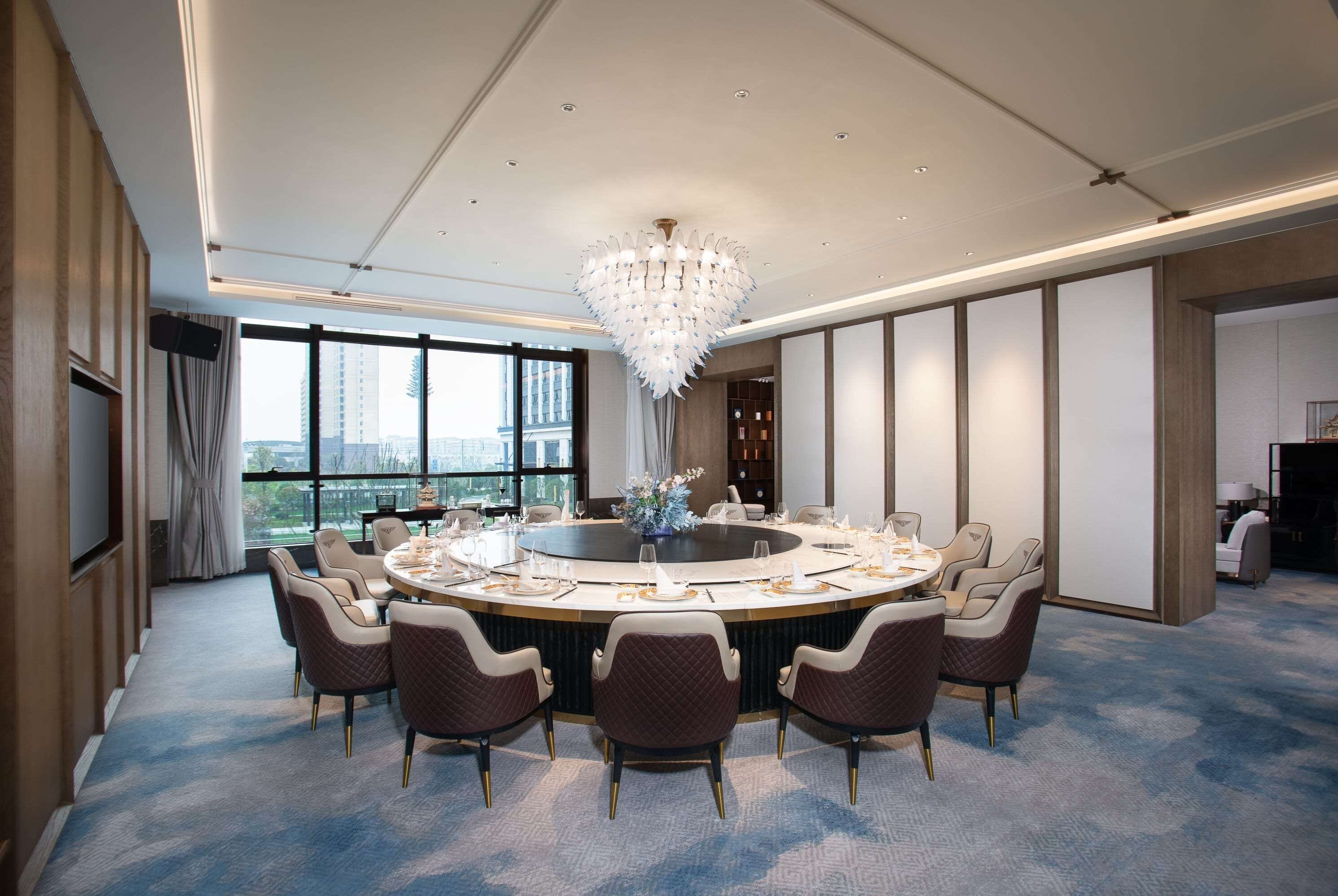 A modern conference room with a round table, comfortable chairs, a chandelier, and large windows offering a city view.