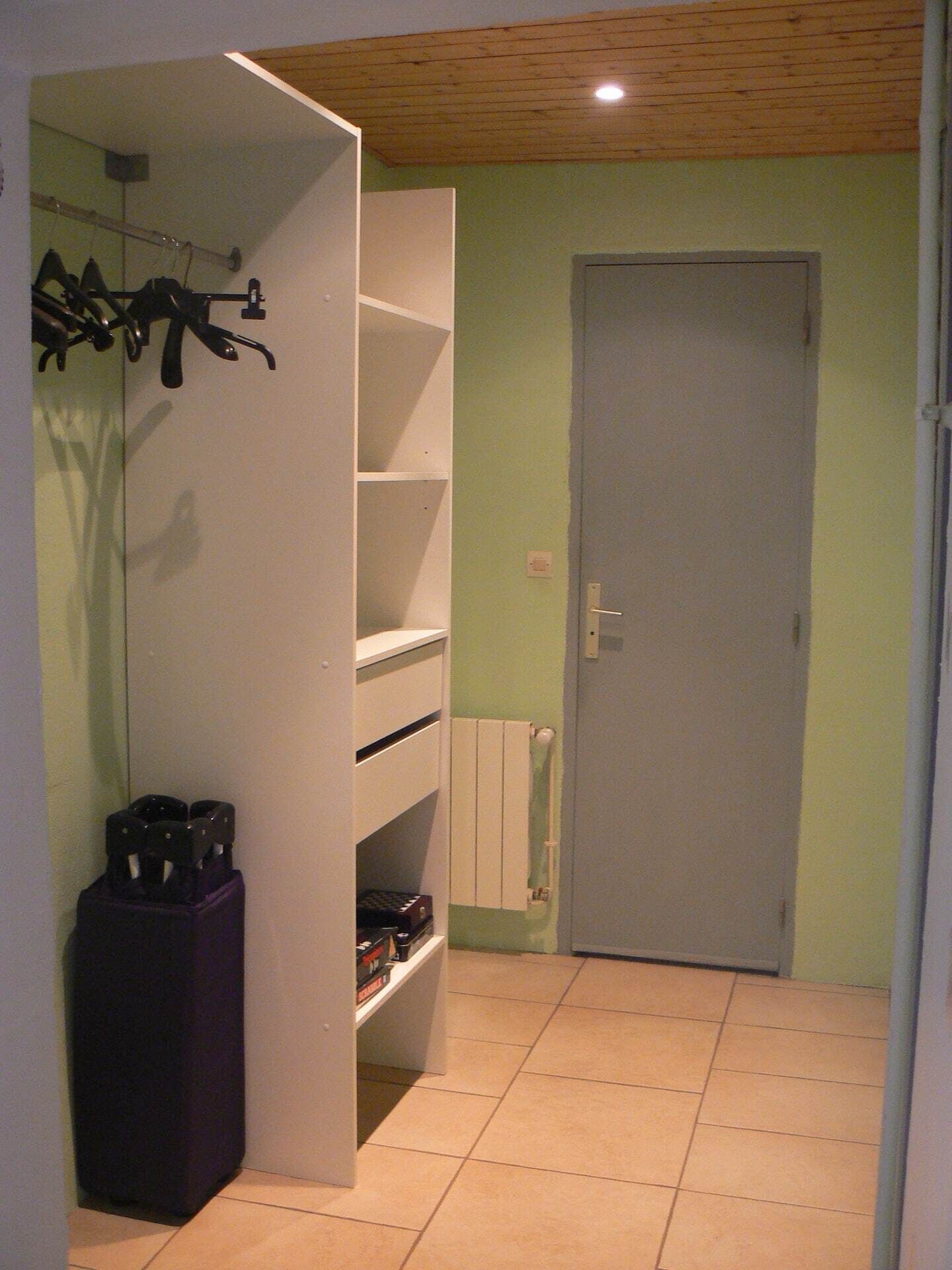 Una habitación pequeña y bien iluminada, con piso de baldosas, un armario blanco con puertas abiertas, una puerta cerrada y un radiador.