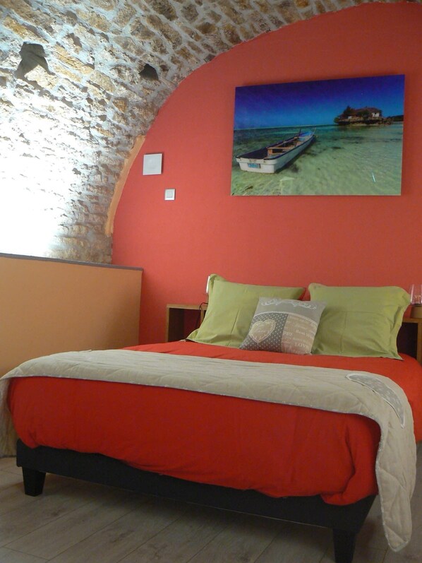 A bedroom with a red bedspread, a wooden headboard, a single bed, and a painting of a boat on a beach.