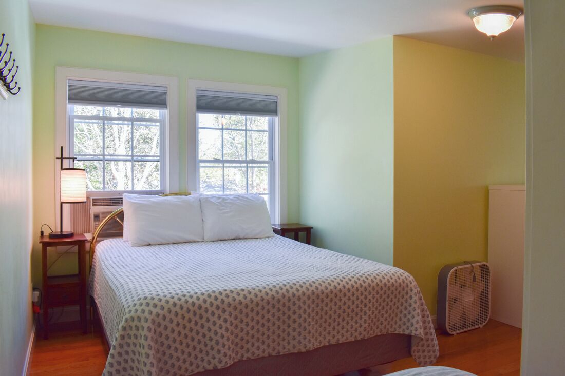 A bedroom with a bed, two windows, a bedside table with a lamp, a radiator, and a standing fan.