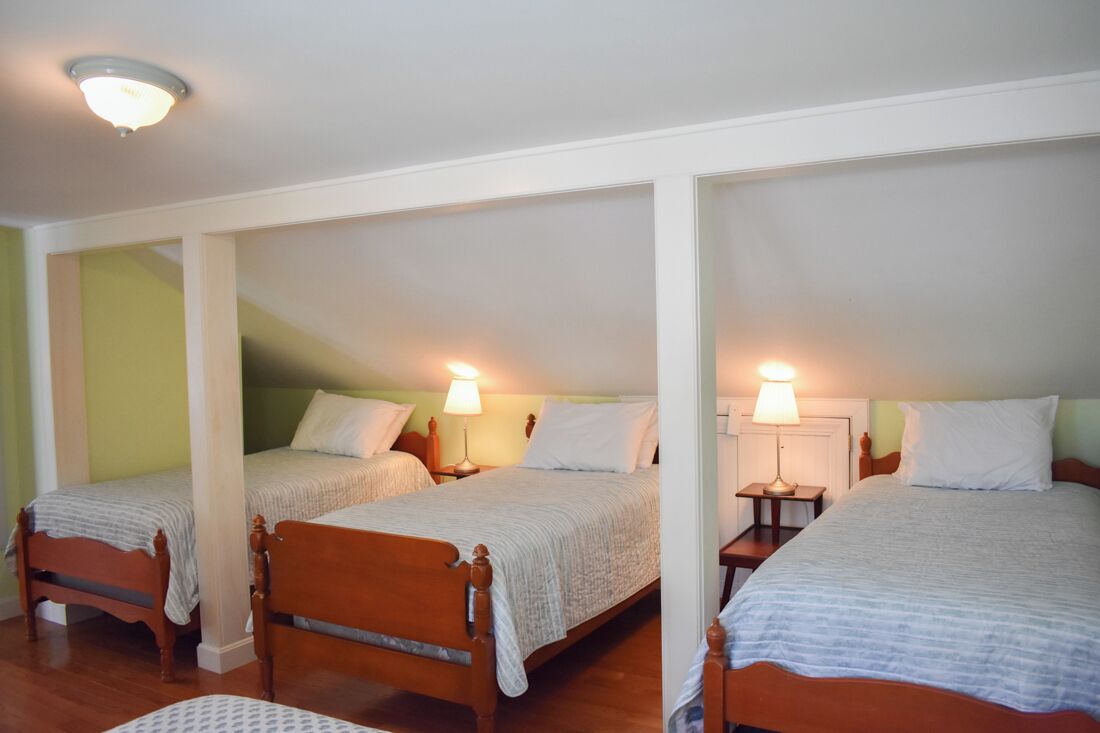 Een kamer met drie houten bedden, elk met een nachtkastje en een lamp. De kamer heeft een schuine kap en een zichtbare lamp.