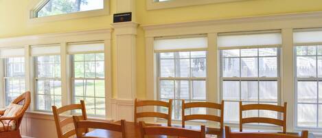 A dining area with a wooden table and chairs, large windows with white shutters, and a view of the outdoors.
