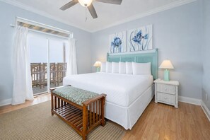 A bedroom with a large bed, a wooden bench, a ceiling fan, a nightstand with a lamp, and artwork on the wall. - 5 Bedroom ~ BEACH HOUSE ~ WEEKLY RENTALS! (Gulf Shores)