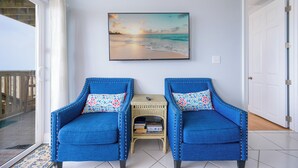 Une pièce avec deux fauteuils bleus, une petite table sur laquelle sont posés des livres, un tableau représentant un coucher de soleil sur l’océan et une porte.