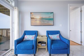 A room with two blue armchairs, a small table with books, a painting of a sunset over the ocean, and a door. - 5 Bedroom ~ BEACH HOUSE ~ WEEKLY RENTALS! (Gulf Shores)