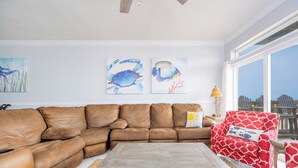 Un salon avec un canapé d’angle en cuir marron, une table basse en bois et un fauteuil à motifs rouges. Deux tableaux représentant des poissons sont accrochés au mur.