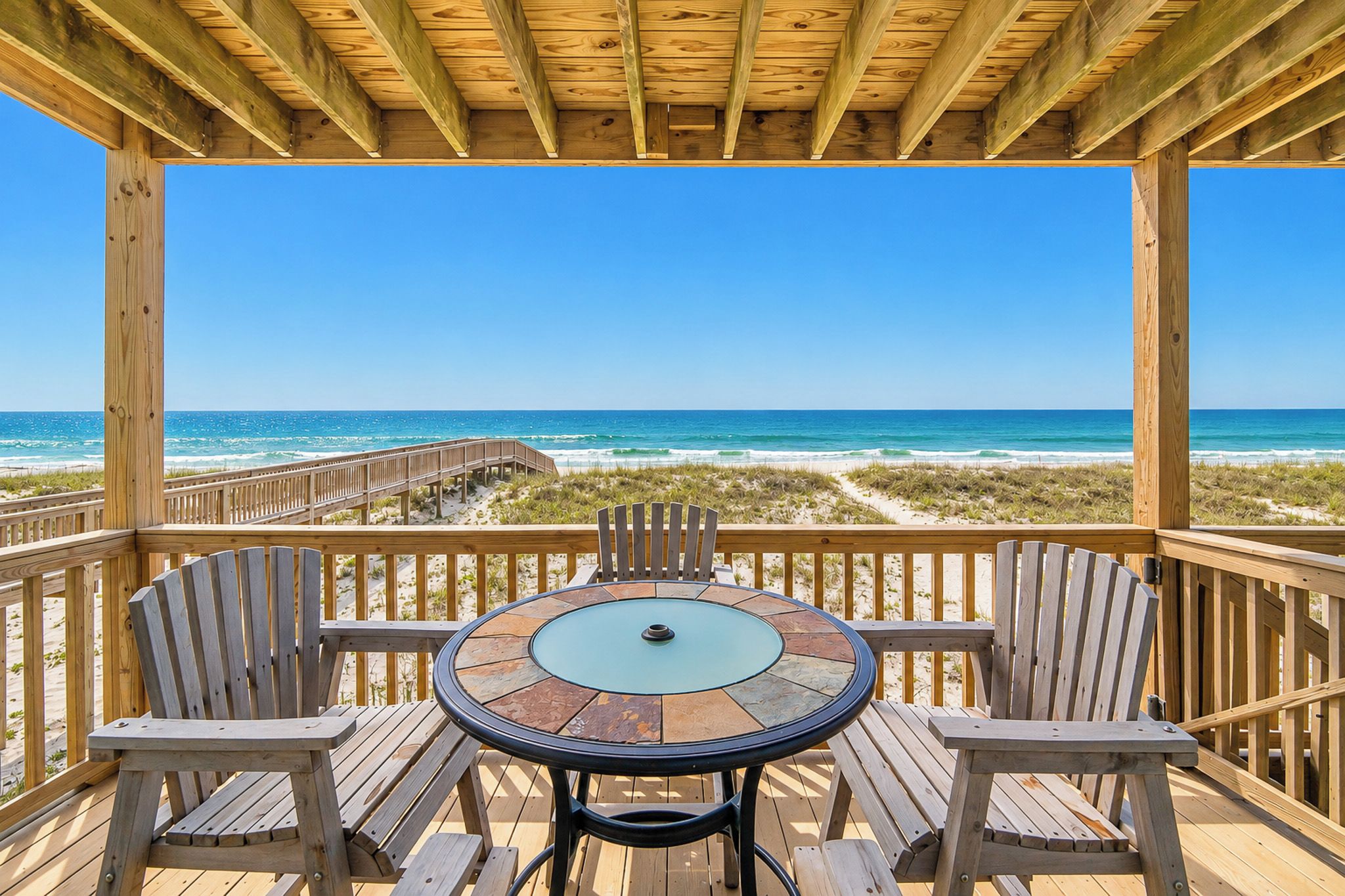 A wooden deck with a round table and chairs, overlooking a beach with a wooden walkway.
