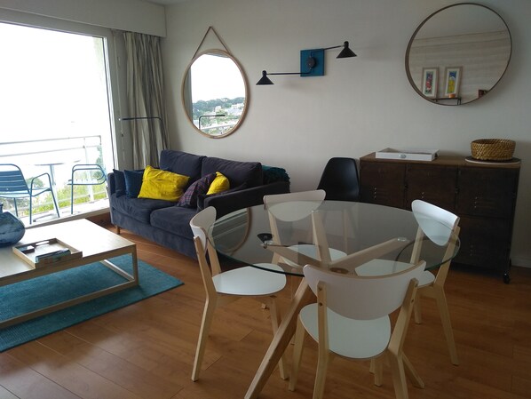 A living room with a sofa, a dining table with chairs, a large mirror, and a view through a window.