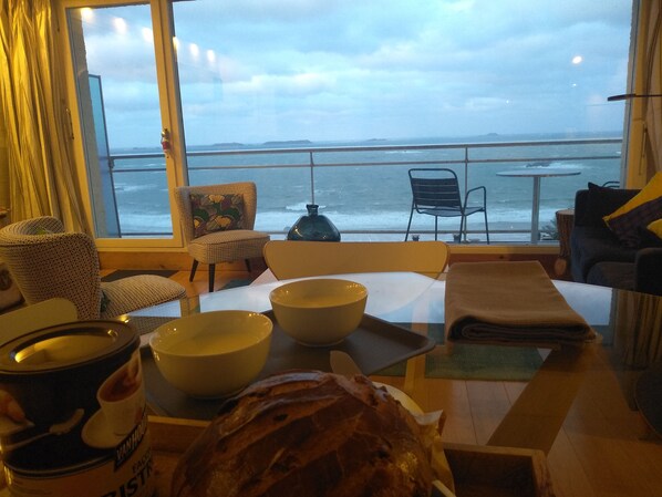 A dining area with a view of the sea, featuring a table with bowls, a loaf of bread, and a can of VanHouten chocolate.