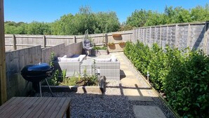Un patio trasero con terraza de madera, una parrilla, mobiliario de mimbre y una zona de jardín.