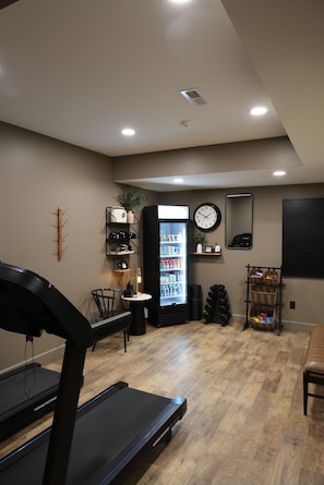 A gym with treadmills, a wall-mounted clock, a refrigerator stocked with beverages, and a mirror. - L3nee's Lay-Up by Creative Cabins (Logan)