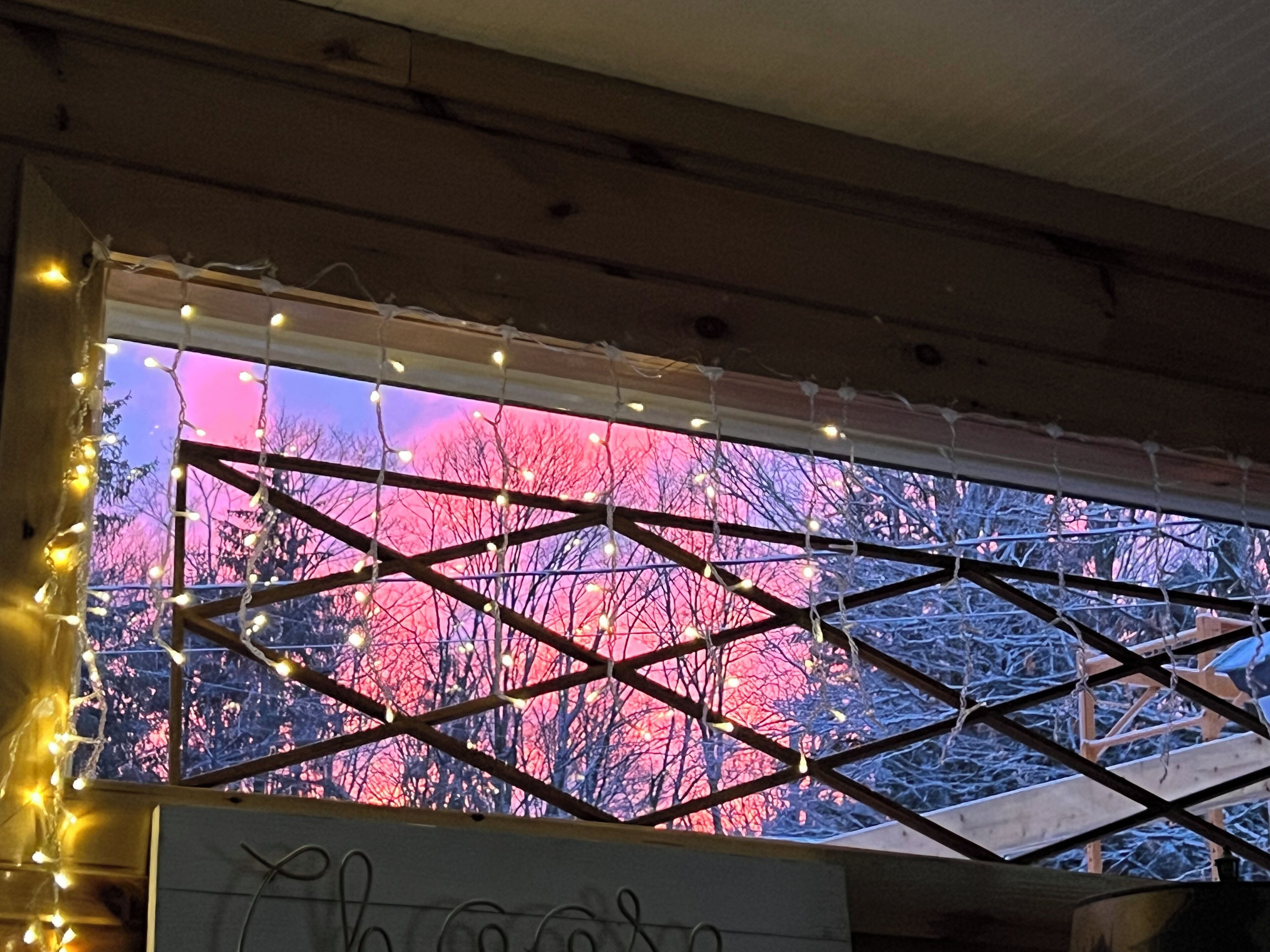 Habitación con ventana decorada con luces de guirnalda y vista a árboles contra un cielo rosado.