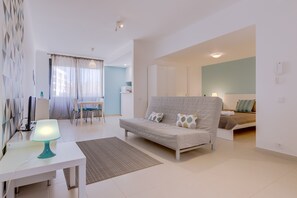Een moderne, goed verlichte woonkamer met een bank, een eethoek en een afzonderlijke slaapkamer met een bed.