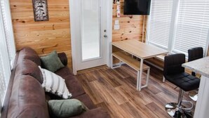 A cozy cabin interior with a wooden ceiling, a ceiling fan, a brown leather couch, a dining table with chairs, and a TV mounted on the wall.