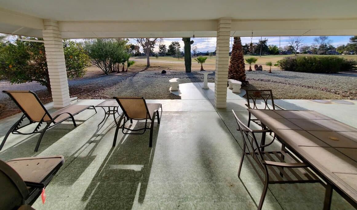 A covered patio area with outdoor seating, including chairs and a table, overlooking a landscaped garden with trees and shrubs.