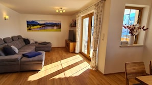 A living room with a sectional sofa, a flat-screen TV, a painting of a landscape, and a window with curtains. - Apartment Hipp No. 3 - 90m² new building on the 1st floor with balcony and mountain view in a top location (Halblech)