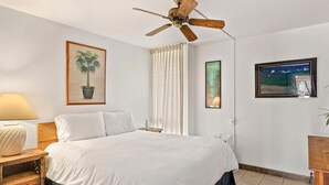 Une chambre d’hôtel avec un lit, un ventilateur de plafond, une fenêtre avec des rideaux, une table de chevet avec une lampe et des œuvres d’art accrochées aux murs.