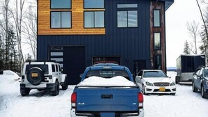 A modern two-story house with a garage, parked cars, and a snow-covered driveway.