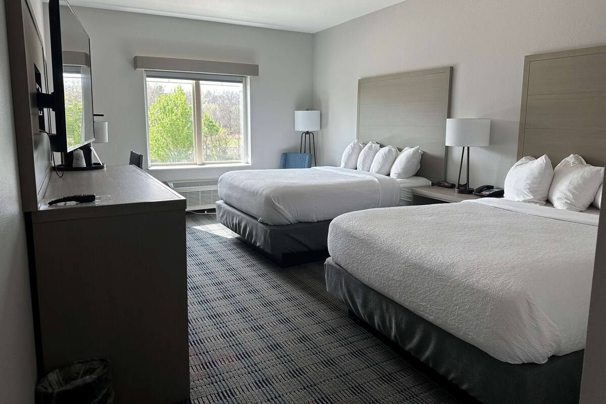 A hotel room with two beds, a desk, a chair, a lamp, and a large window.