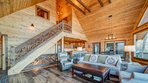 Ruim houten interieur van een chalet met een trap, een zithoek met banken en een salontafel, en een keuken op de achtergrond.