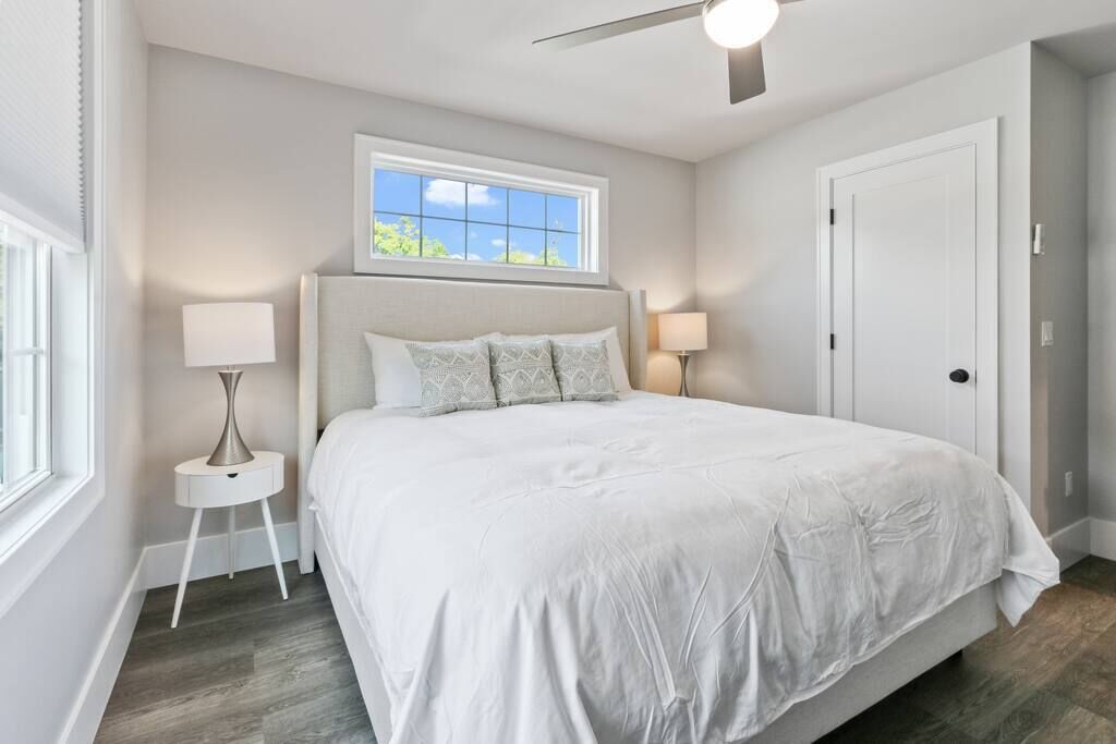 A bedroom with a bed, two bedside tables with lamps, a ceiling fan, a window, and a door.