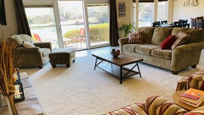 A living room with a sofa, ottoman, coffee table, and a view of a lake through large glass doors. - Spoil yourself with this beautiful Lake Balboa retreat (Hot Springs Village)