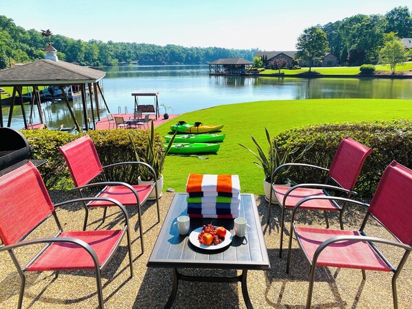 A lakeside patio with red chairs, a table with a plate of fruit and a mug, and kayaks in the background.