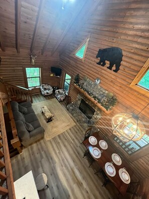 A log cabin interior with a stone fireplace, a sofa, a dining table with plates, and a bear decoration on the ceiling.