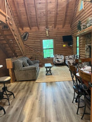 A log cabin interior with a sofa, dining table, chairs, a small table, and a staircase.