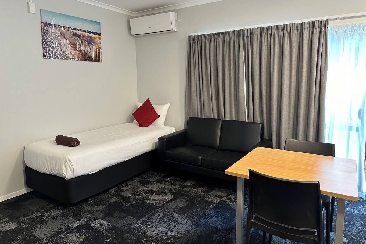 A compact room with a bed, a sofa, a table, and a chair. There is a painting on the wall and an air conditioning unit mounted above the sofa.