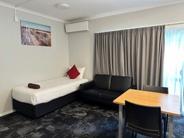 Una habitación compacta con cama, sofá, mesa y silla. Hay un cuadro en la pared y un equipo de aire acondicionado sobre el sofá.