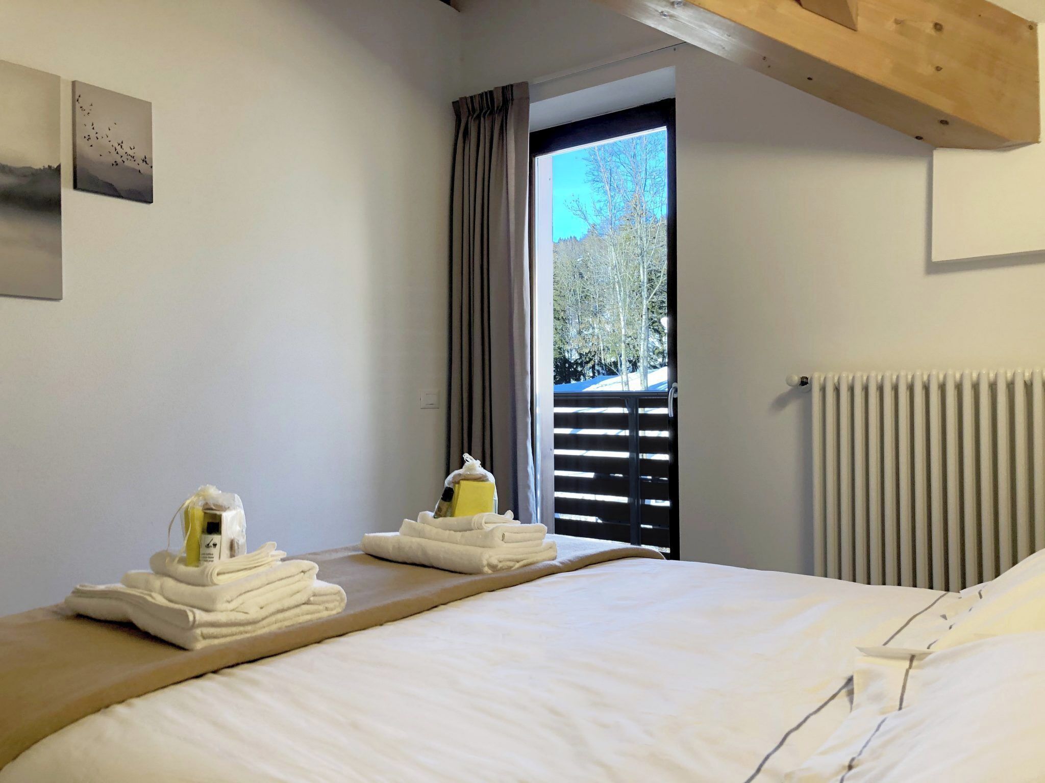 Una camera d'albergo con un letto, una testiera in legno, una finestra con tende e termosifone, e vista su alberi.