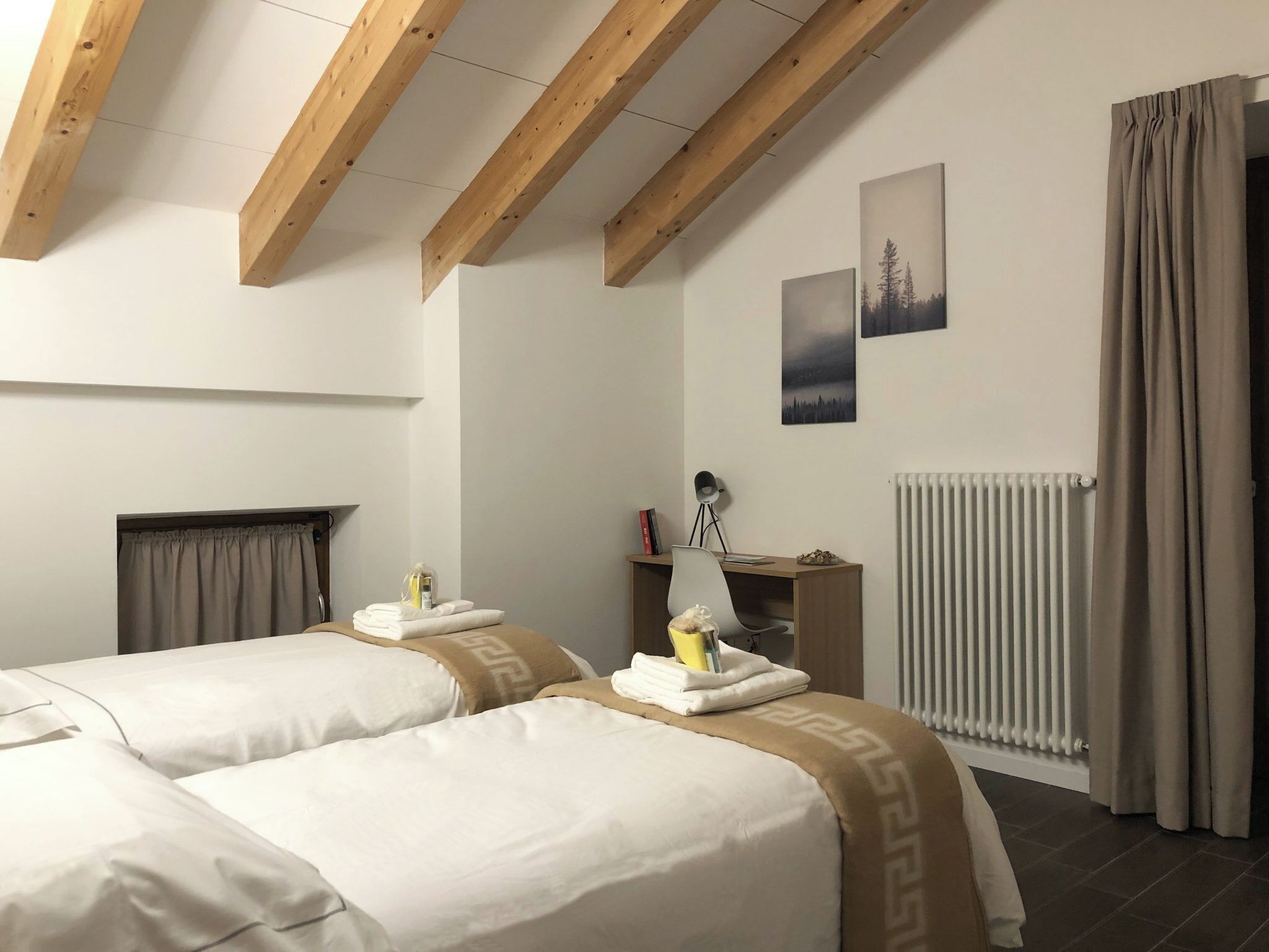A hotel room with two beds, a desk with a chair, a radiator, and a window with curtains.