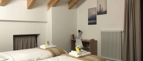 A hotel room with two beds, a desk with a chair, a radiator, and a window with curtains.