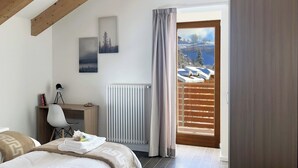 Ein Schlafzimmer mit Bett, Schreibtisch, Stuhl, Heizkörper und einem Fenster mit Blick auf schneebedeckte Dächer.