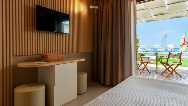 A modern hotel room with a large bed, a flat-screen TV mounted on a wooden panel wall, a small desk with a chair, and a view of a beach with umbrellas and sailboats. - Hotel Il Tempio (Casal Velino)