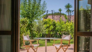 A room with a bed, two wooden chairs, a small table, and a view of a garden with trees and a building in the background. - Hotel Il Tempio (Casal Velino)
